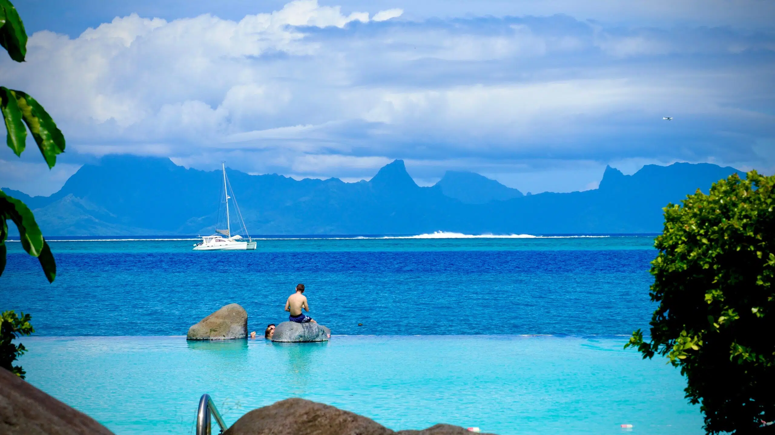 A person sitting on the edge of a pool in Tahiti with a sailboat and mountains in the distance.